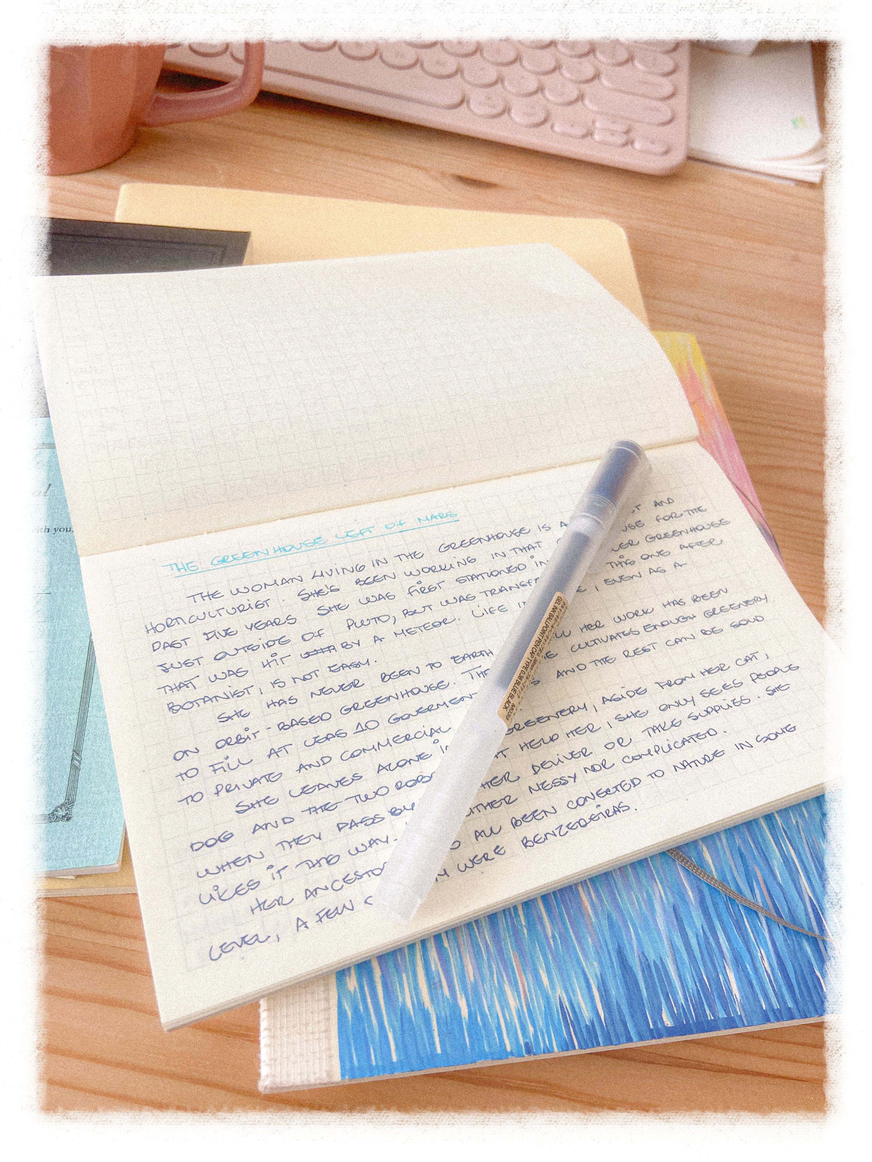 A small notebook is open over a wooden table. The paper in the notebook has a light blue grid and text is handwritten on it. Underneath the notebook, a colourful sketchbook.